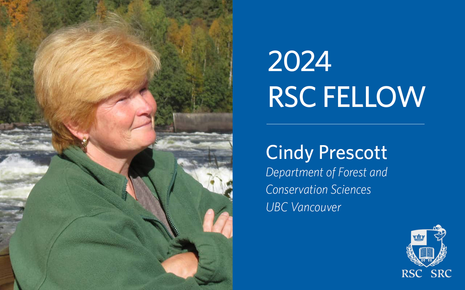 Cindy Prescott has medium length strawberry blonde hair and is wearing gold hoop earrings. She is wearing a green pullover sweater and is facing profile to the camera, smiling at an unknown subject in the distance. There appears to be a rushing river and autumnal forest behind her.