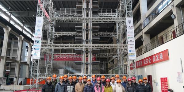A group pof about 20 people in red and orange hard hats stand in front of a multi-level earthquake simulator