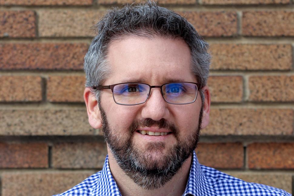 Corey Stephenson has short, salt-and-pepper hair and a dark beard, and wears square glasses. He smiles softly to the camera.