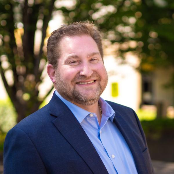 2025 Headshot of Leslie Grad: a man with light brown hair and a short beard, wearing a blue dress shirt and navy blue blazer